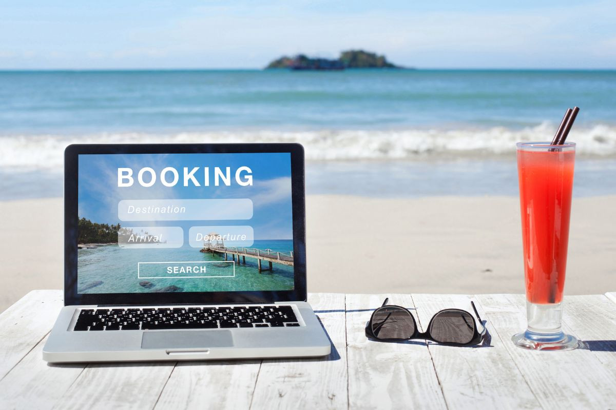 A laptop showing a booking screen is placed on a beachside table next to a pair of sunglasses and a tall, refreshing drink, with the ocean in the background.